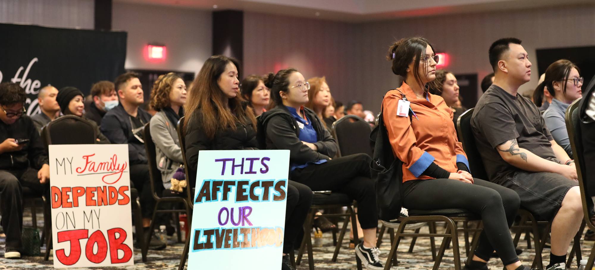 Cardroom employees seated at a public hearing, holding handmade signs reading "My family depends on my job" and "This affects our livelihood".